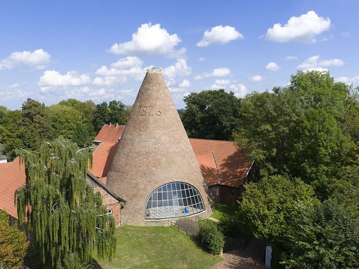 Historischer kegelförmiger Tafelturm der Glashütte Gernheim mit Backsteinfassade und großem Fenster, umgeben von roten Dächern und Bäumen.