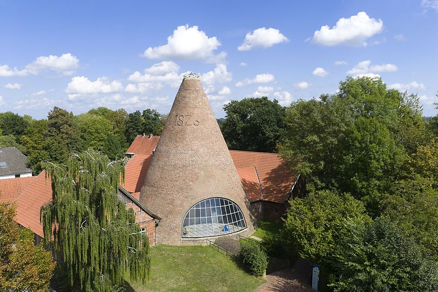 Historischer kegelförmiger Tafelturm der Glashütte Gernheim mit Backsteinfassade und großem Fenster, umgeben von roten Dächern und Bäumen.